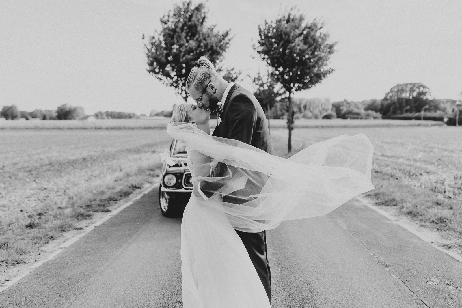 Schwarz Weiß Foto von Brautpaar, dass sich vor altem Ford Mustang küsst