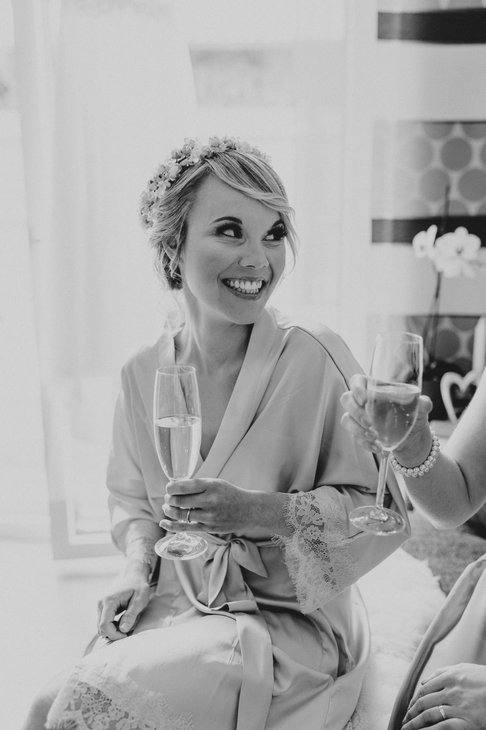 Ein schwarz weiß Foto einer Braut, die einen Kimono beim Getting Ready trägt, ein Glas Sekt in der Hand hält und eine andere Person anlächelt, die nicht zu sehen ist.